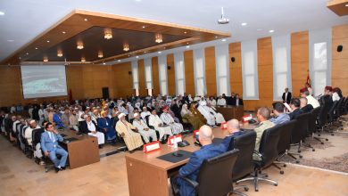 Audience seated in a modern conference hall listening to a panel at the front, with a projector screen on the side and multiple flags nearby.