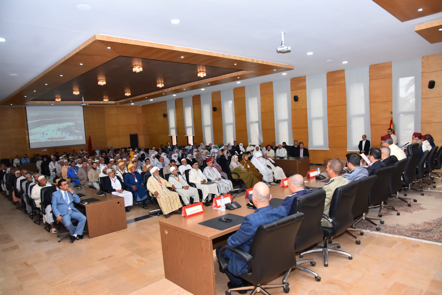 Audience seated in a modern conference hall listening to a panel at the front, with a projector screen on the side and multiple flags nearby.