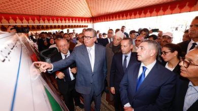 Group of dignitaries in suits under a red patterned tent, a man in a gray suit points to a display vehicle as others observe.