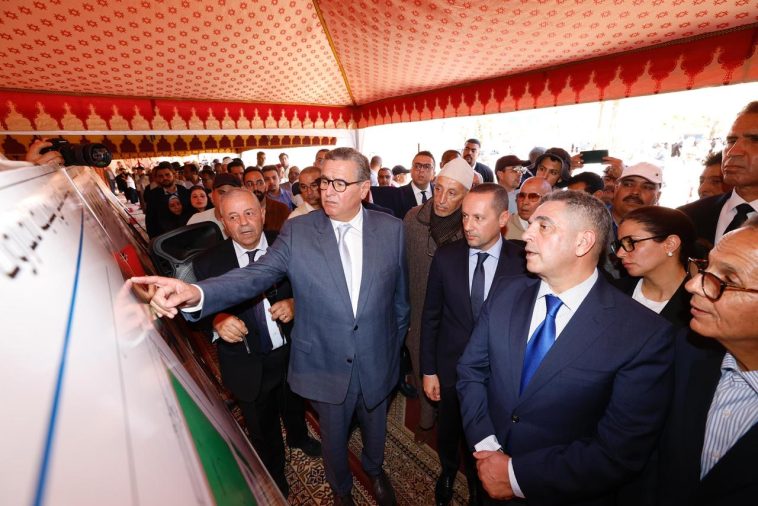 Group of dignitaries in suits under a red patterned tent, a man in a gray suit points to a display vehicle as others observe.