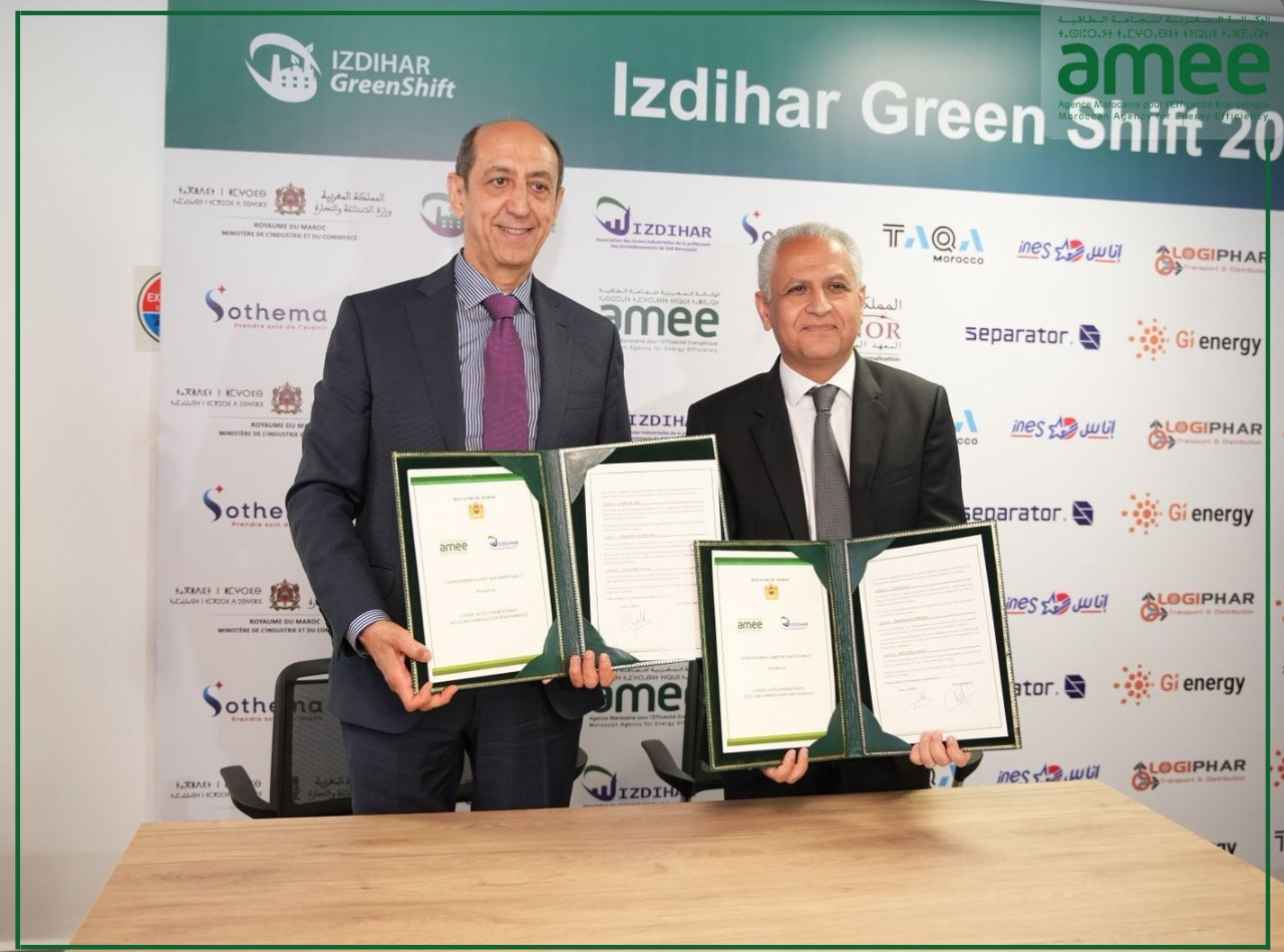 Two men in suits hold framed certificates during a signing ceremony in front of a branded backdrop.