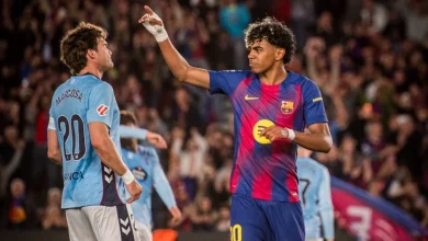 Barcelona player wearing number 10 in blue-and-red kit raises his arm while facing an opposing player in light-blue jersey; cheering crowd in the stands behind them.
