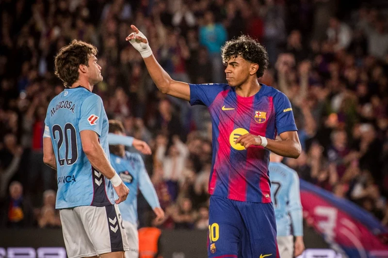 Barcelona player wearing number 10 in blue-and-red kit raises his arm while facing an opposing player in light-blue jersey; cheering crowd in the stands behind them.