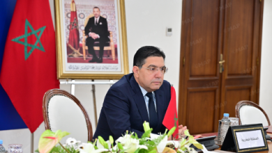 Moroccan official seated at a conference table with a flag and framed portrait in the background.