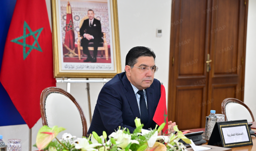Moroccan official seated at a conference table with a flag and framed portrait in the background.