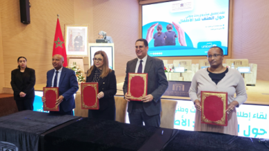 Four professionals stand at a ceremony holding red-bound certificates on a stage, with a Moroccan flag in the background.
