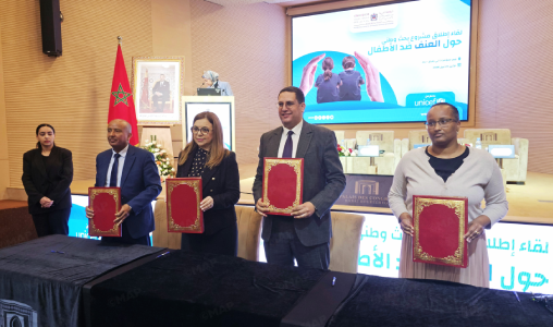 Four professionals stand at a ceremony holding red-bound certificates on a stage, with a Moroccan flag in the background.