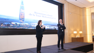 Two presenters on a stage in front of a large screen displaying a French welcome message, ‘Bienvenue à bord de la Tour Mohammed VI’ behind them.