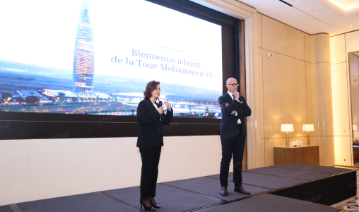 Two presenters on a stage in front of a large screen displaying a French welcome message, ‘Bienvenue à bord de la Tour Mohammed VI’ behind them.