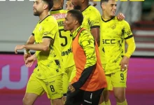 Four soccer players in neon yellow uniforms celebrate on the field, one in an orange bib in front of the group