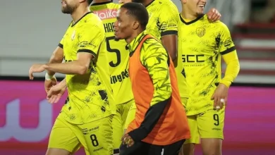 Four soccer players in neon yellow uniforms celebrate on the field, one in an orange bib in front of the group