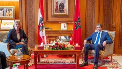 Two officials in formal suits sit across from each other in a wood-paneled room with Austrian and Moroccan flags and a flower arrangement on the table behind them.