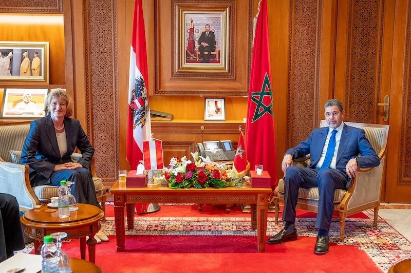 Two officials in formal suits sit across from each other in a wood-paneled room with Austrian and Moroccan flags and a flower arrangement on the table behind them.