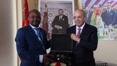 Two men in suits exchange a black presentation box during a formal meeting, with flags and portraits in the background.