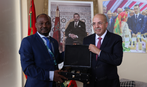 Two men in suits exchange a black presentation box during a formal meeting, with flags and portraits in the background.