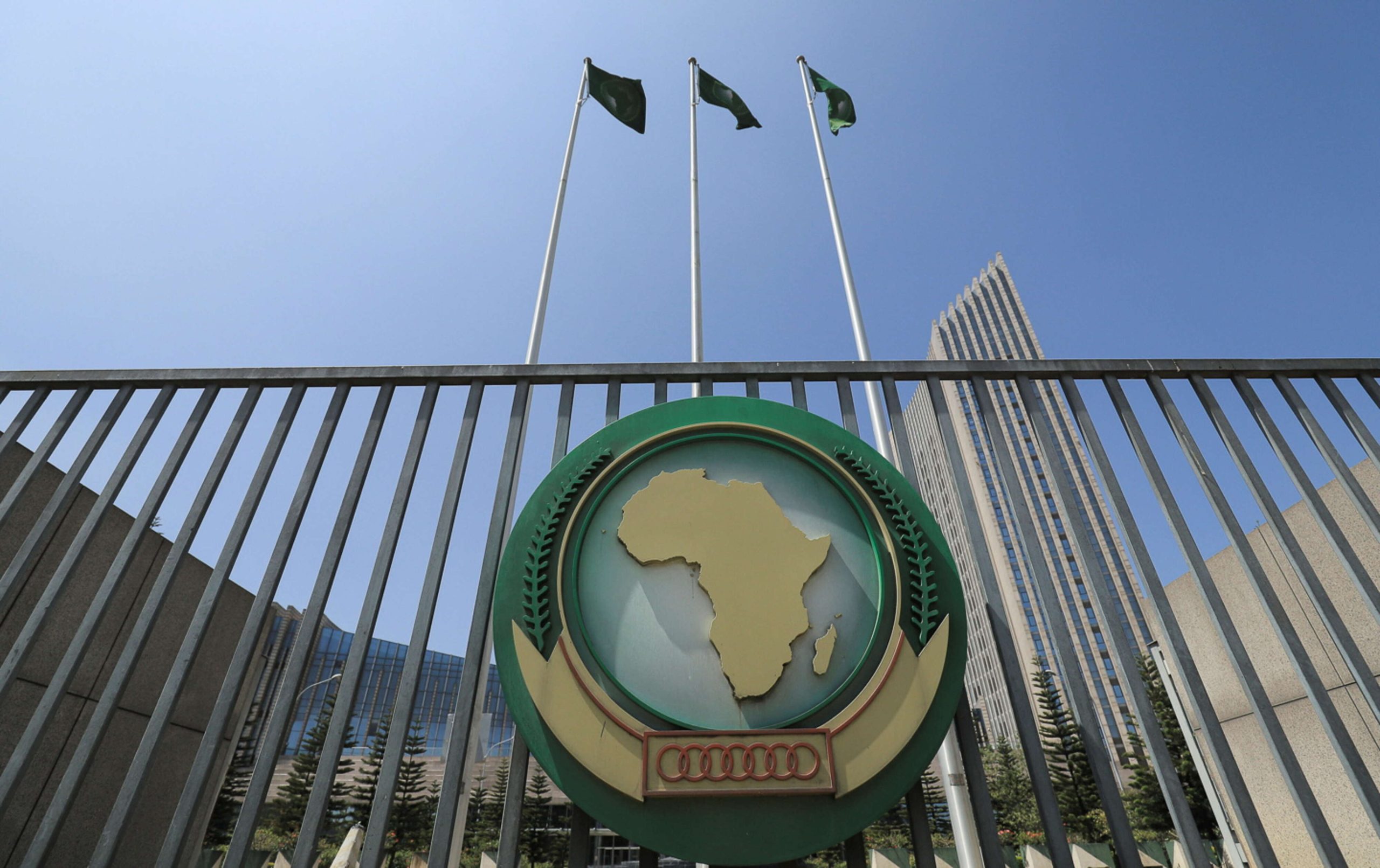 Circular emblem showing a gold map of Africa on a green field, mounted on a gate with a railing, flanked by flagpoles and a tall building against a blue sky