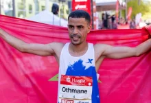 Male runner crosses the finish line, arms outstretched, tearing through a red banner at an outdoor race event.