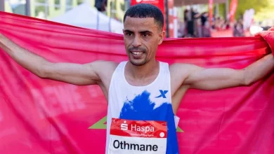 Male runner crosses the finish line, arms outstretched, tearing through a red banner at an outdoor race event.
