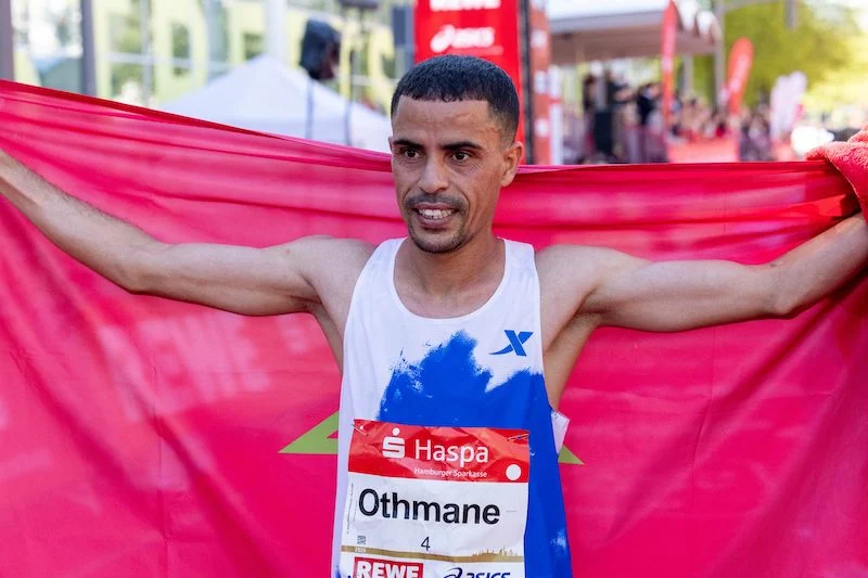 Male runner crosses the finish line, arms outstretched, tearing through a red banner at an outdoor race event.