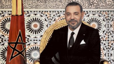 Man in a black suit sits on a gold throne beside a Moroccan flag, with an intricate tiled backdrop.