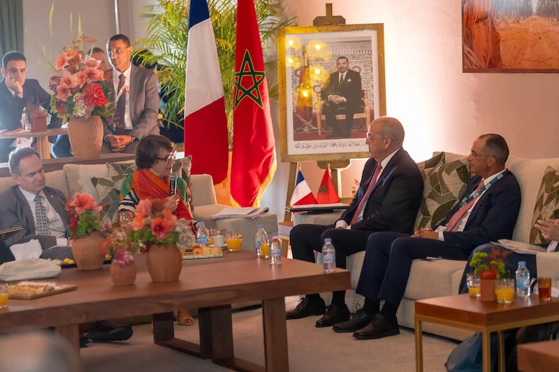 Diplomatic meeting in a living-room style room with flags, a large portrait, and officials seated on sofas around a coffee table with flowers.