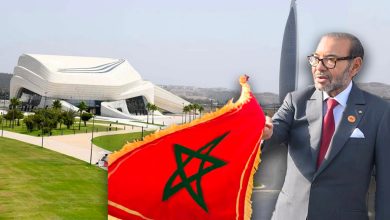 Modern white sculptural building with green lawn; a suited man holds a Moroccan flag in the foreground on the right.
