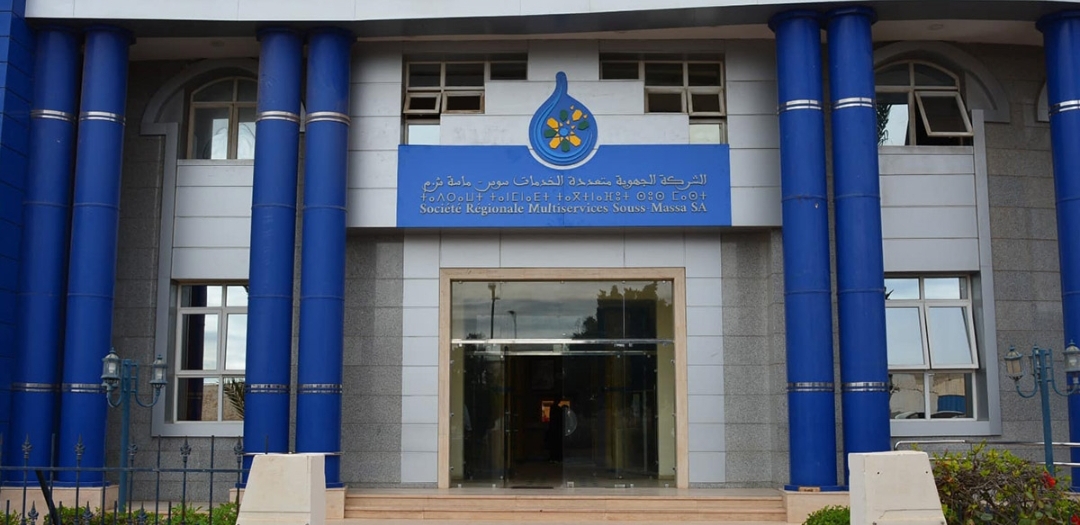 Front façade of a blue-columned public building with a glass entrance and a blue sign above the door written in Arabic and French style text.