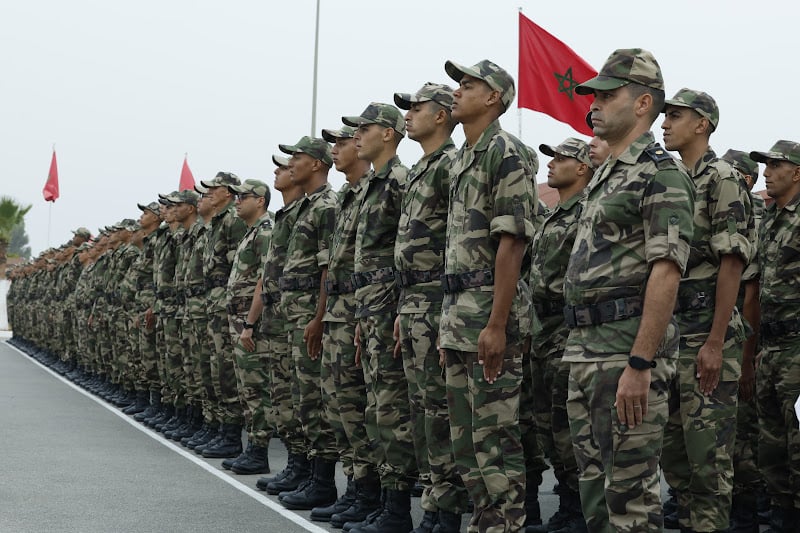 Line of soldiers in camouflage uniforms standing at attention during a parade, Moroccan flag in the background.