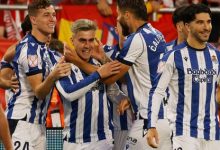 Soccer players in blue-and-white striped kits celebrate a goal, hugging and smiling on the field with red-clad fans in the stands.