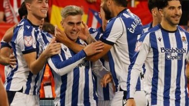 Soccer players in blue-and-white striped kits celebrate a goal, hugging and smiling on the field with red-clad fans in the stands.