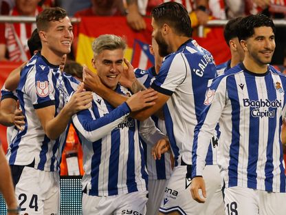 Soccer players in blue-and-white striped kits celebrate a goal, hugging and smiling on the field with red-clad fans in the stands.
