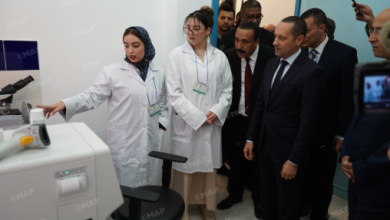 Two female scientists in white lab coats demonstrate a device to a group of suited officials in a lab, pointing at equipment on a white bench.