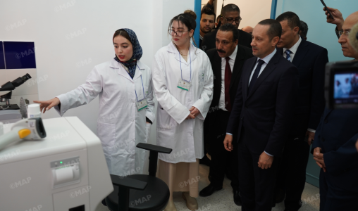 Two female scientists in white lab coats demonstrate a device to a group of suited officials in a lab, pointing at equipment on a white bench.
