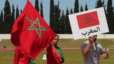 Moroccan flag waving on a sports field with people nearby, sign shows Morocco in Arabic (المغرب).