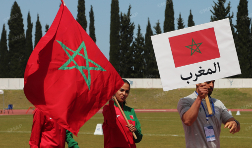 Moroccan flag waving on a sports field with people nearby, sign shows Morocco in Arabic (المغرب).
