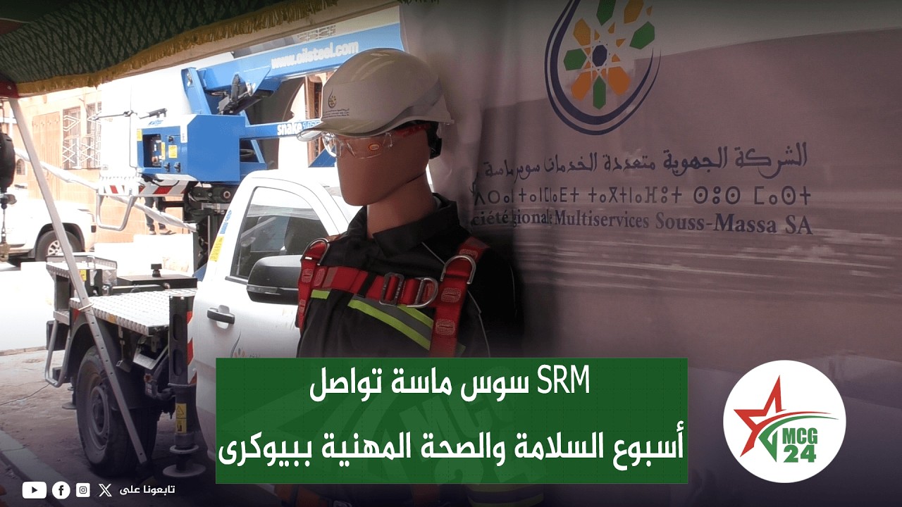 Mannequin wearing a hard hat and safety harness in front of a banner for occupational safety week, with a truck-mounted lift in the background and an MCG 24 logo on the right.