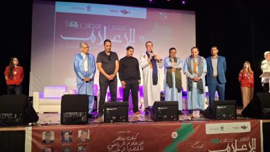 Group of men and women in traditional and formal attire standing on a stage during an event, with a large banner and speakers in the background.