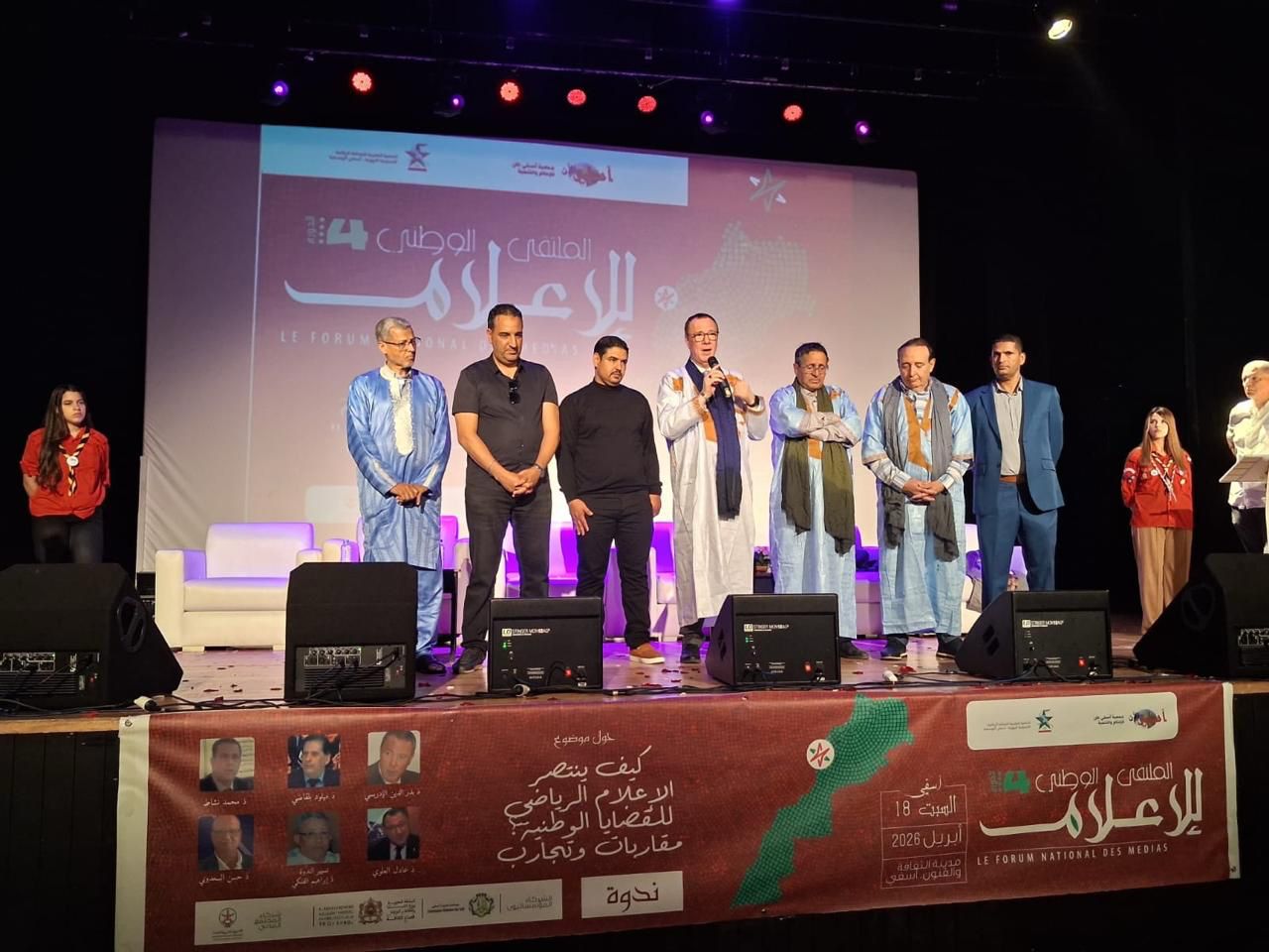 Group of men and women in traditional and formal attire standing on a stage during an event, with a large banner and speakers in the background.