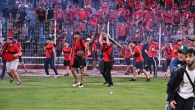 Crowd in red fills the stands as players celebrate on the field, with a man raising a trophy overhead.