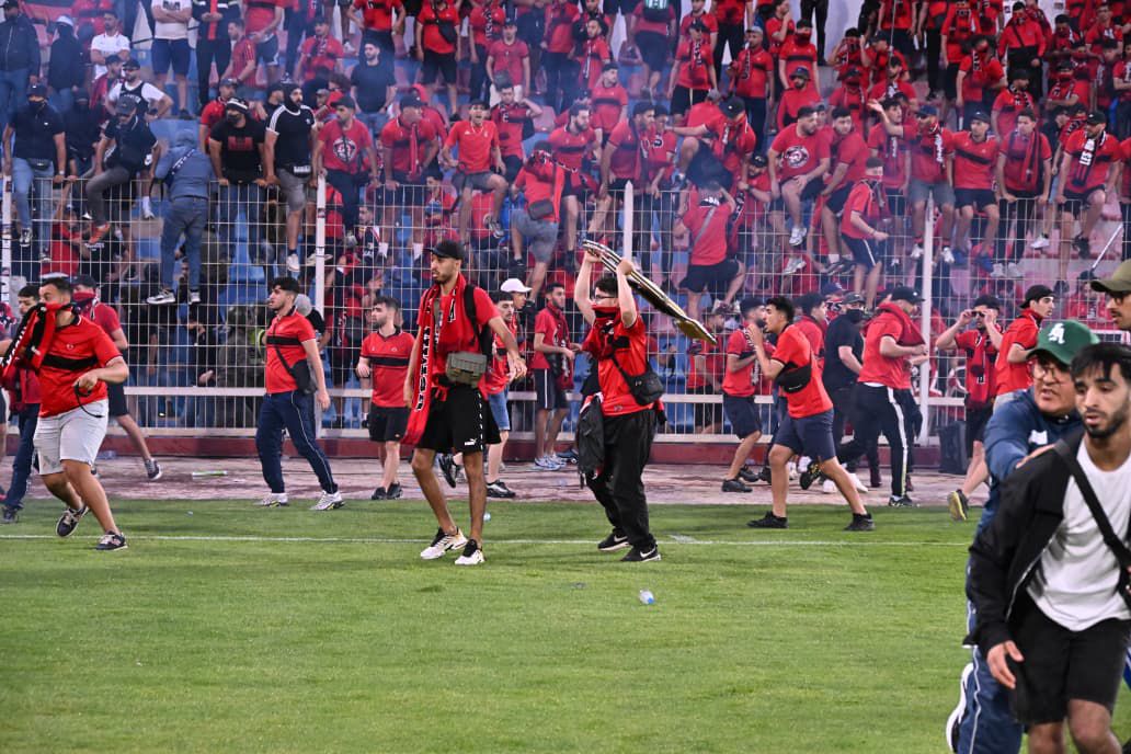 Crowd in red fills the stands as players celebrate on the field, with a man raising a trophy overhead.