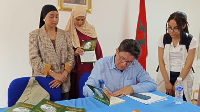 Author seated at a blue table signs a green-covered book while three women stand nearby, Moroccan flag in the background, books on the table.
