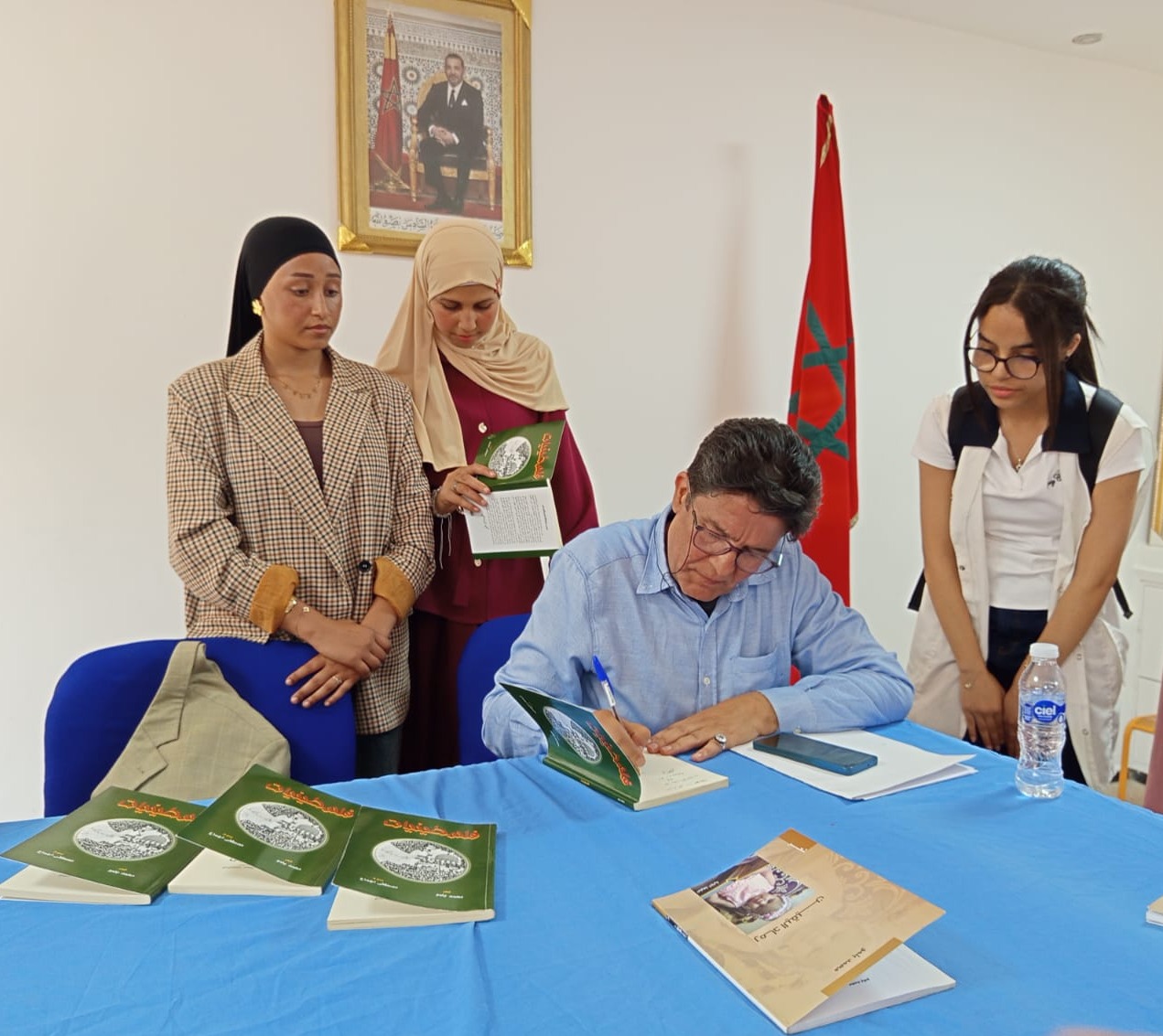 Author seated at a blue table signs a green-covered book while three women stand nearby, Moroccan flag in the background, books on the table.