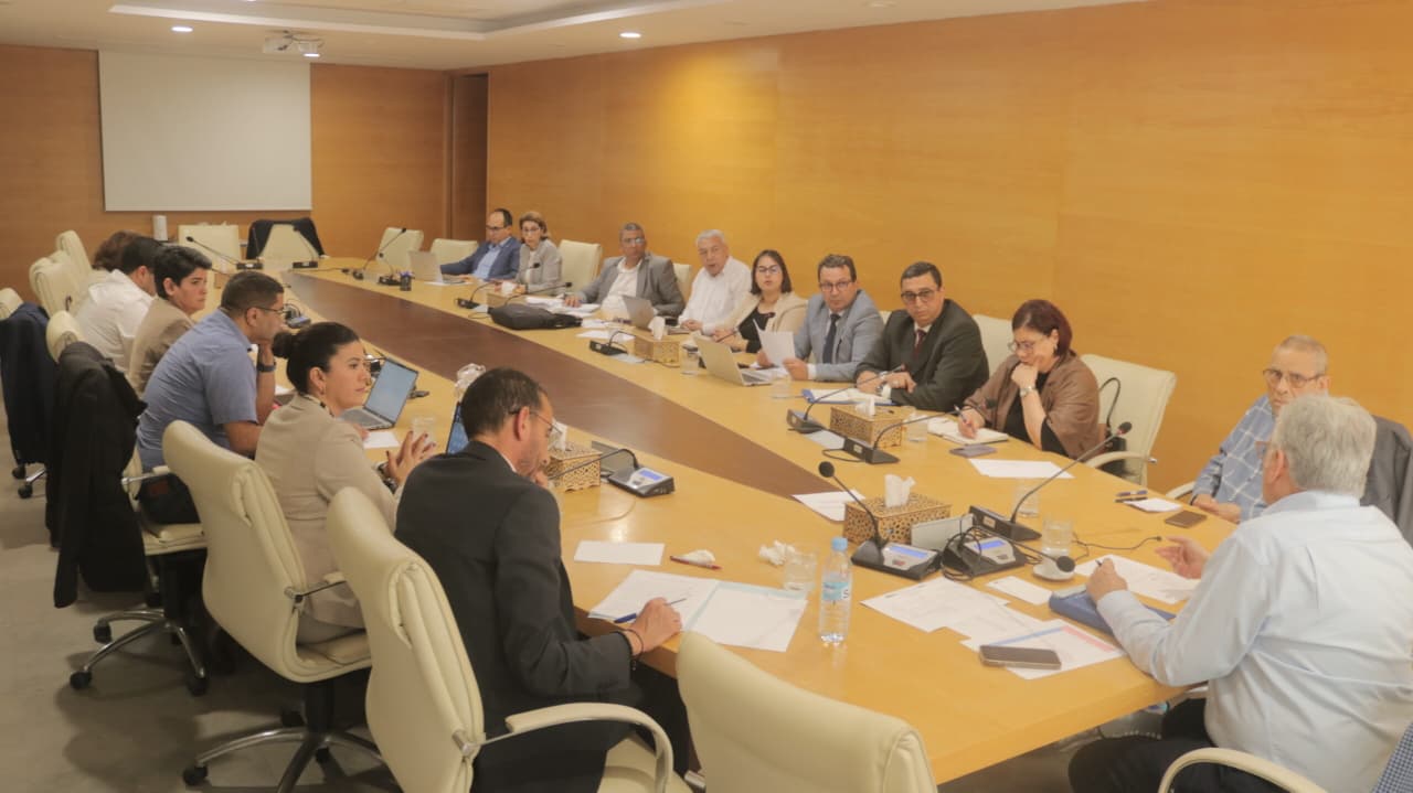 Group of professionals seated around a large conference table in a meeting room, discussing with laptops and documents on the table.