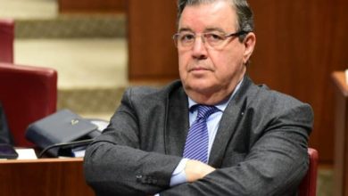 Older man in a gray suit and blue tie sits with arms crossed in a formal meeting room close to the camera.