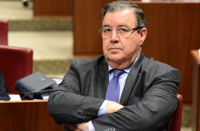 Older man in a gray suit and blue tie sits with arms crossed in a formal meeting room close to the camera.