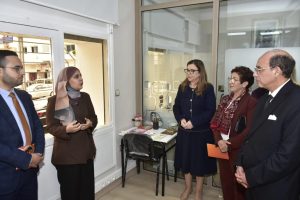 Five professionals in business attire standing in a small office, engaged in a discussion near a desk.