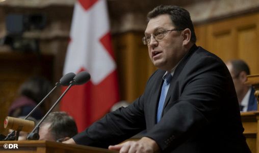 Man in a suit speaking at a wooden podium in a formal government chamber with a Georgian flag behind him.