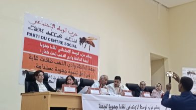 Panel of seven people seated at a conference table on a stage, with a large Arabic banner behind them and a man filming with a smartphone.