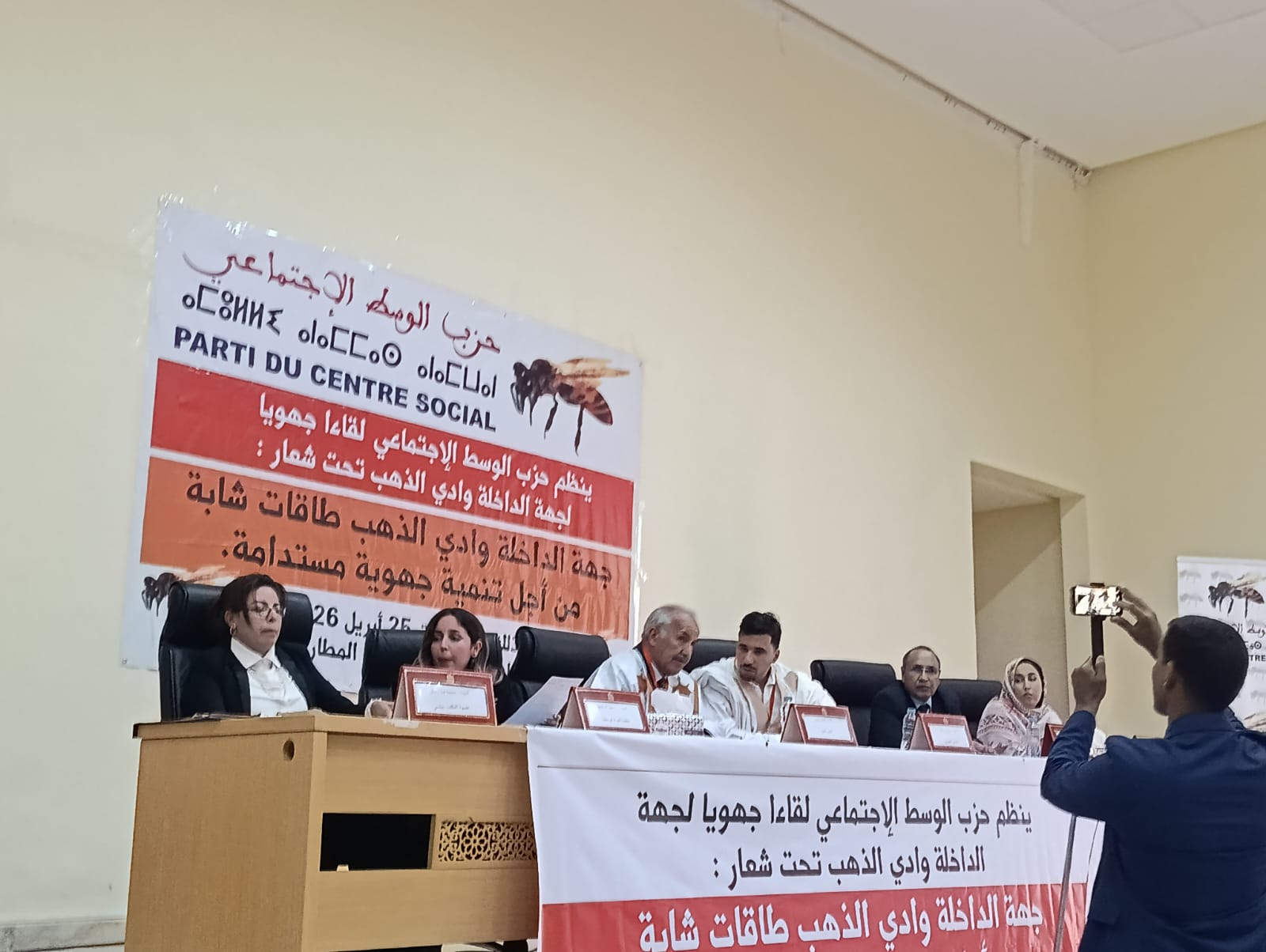 Panel of seven people seated at a conference table on a stage, with a large Arabic banner behind them and a man filming with a smartphone.