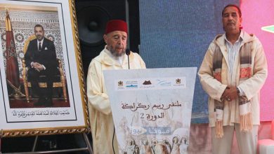 Moroccan man in a red fez speaks at a podium during a cultural event, with a framed portrait of a king on the left and banners nearby.